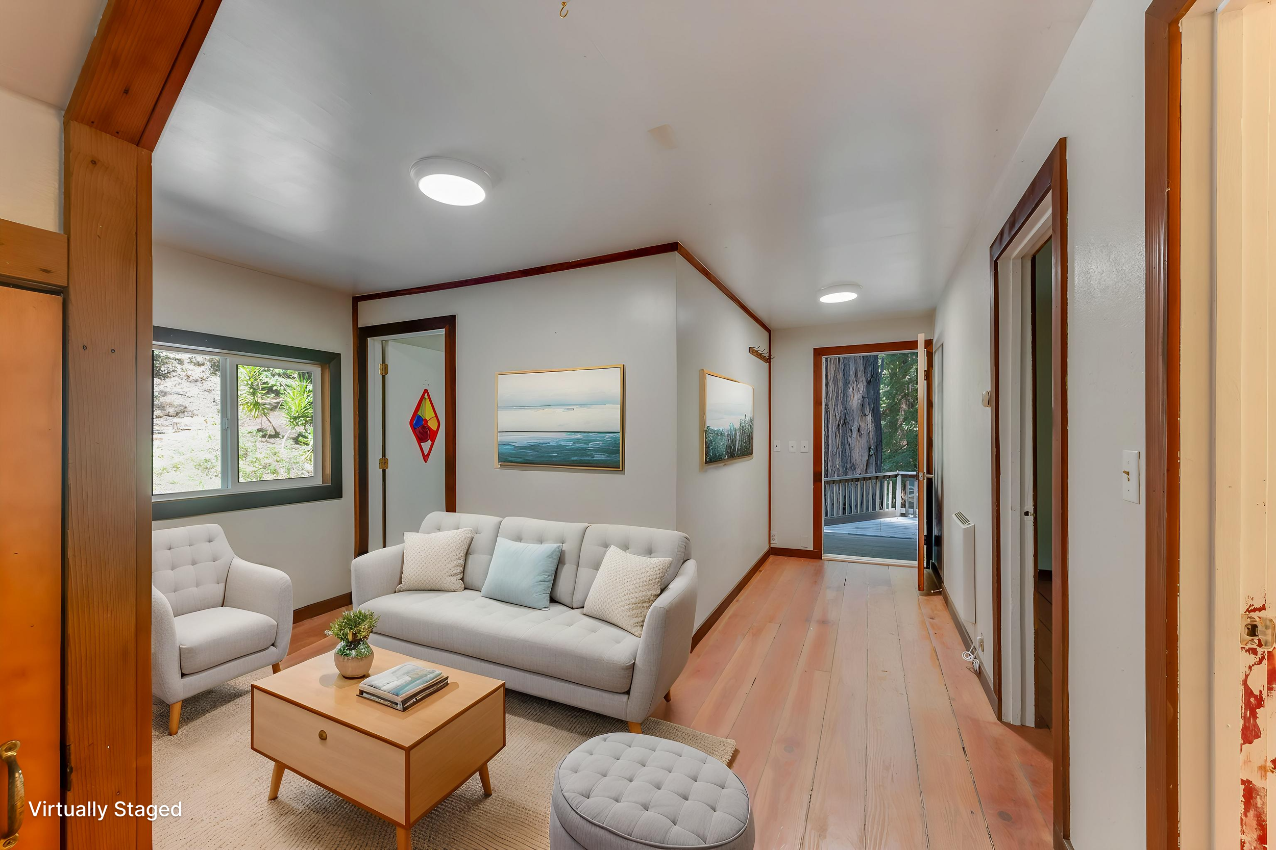 Living room with light wood flooring, white walls, and a white sofa with pillows. There are two white armchairs, a small wooden coffee table with books and a plant. Two paintings hang on the wall. A door opens to a deck outside, and a window shows gr