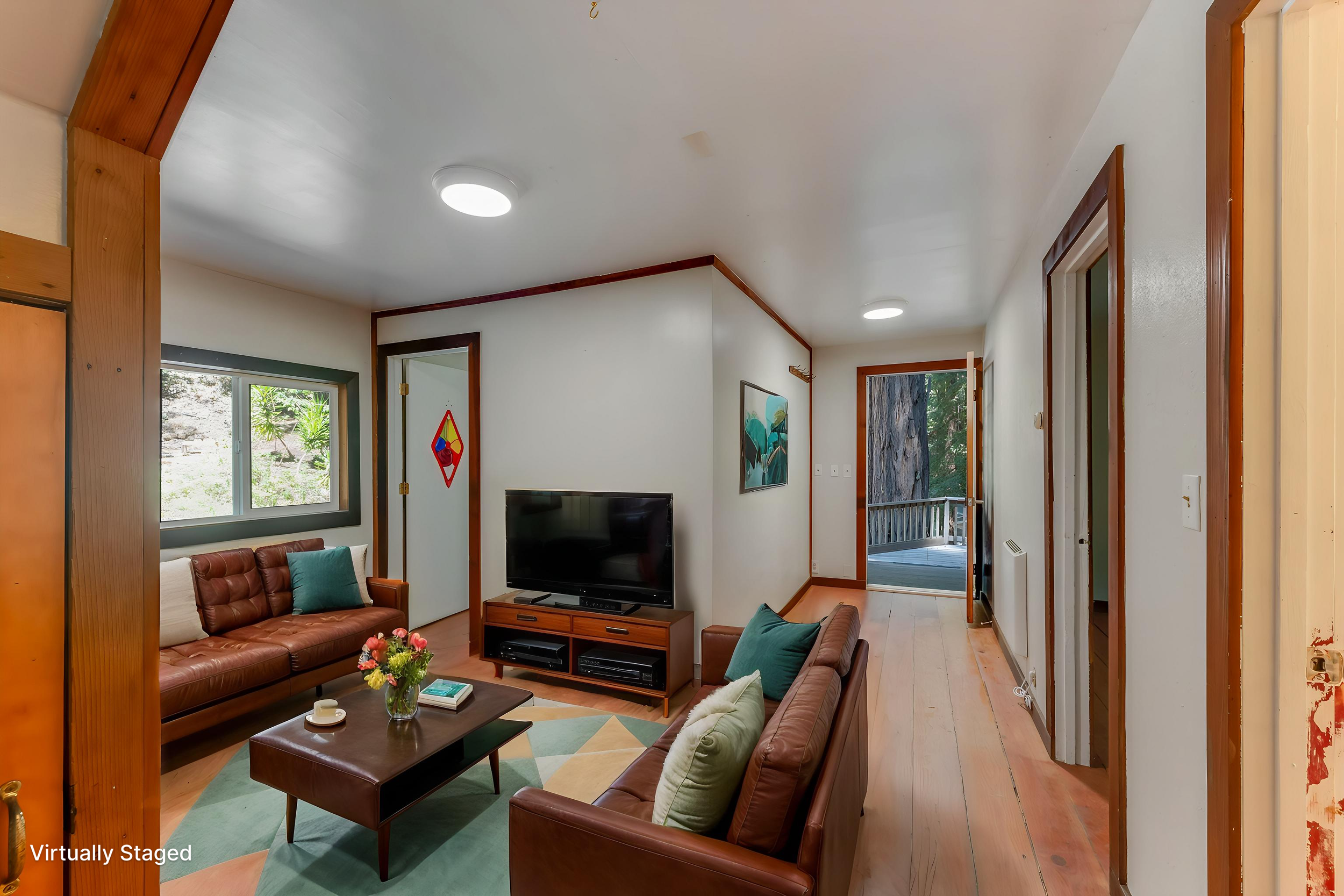 Living room with brown leather sofas, a wooden TV stand with a large flat-screen TV, green pillows on the sofas, a coffee table with a vase of flowers, windows with greenery outside, and a sliding door leading to an outdoor area.
