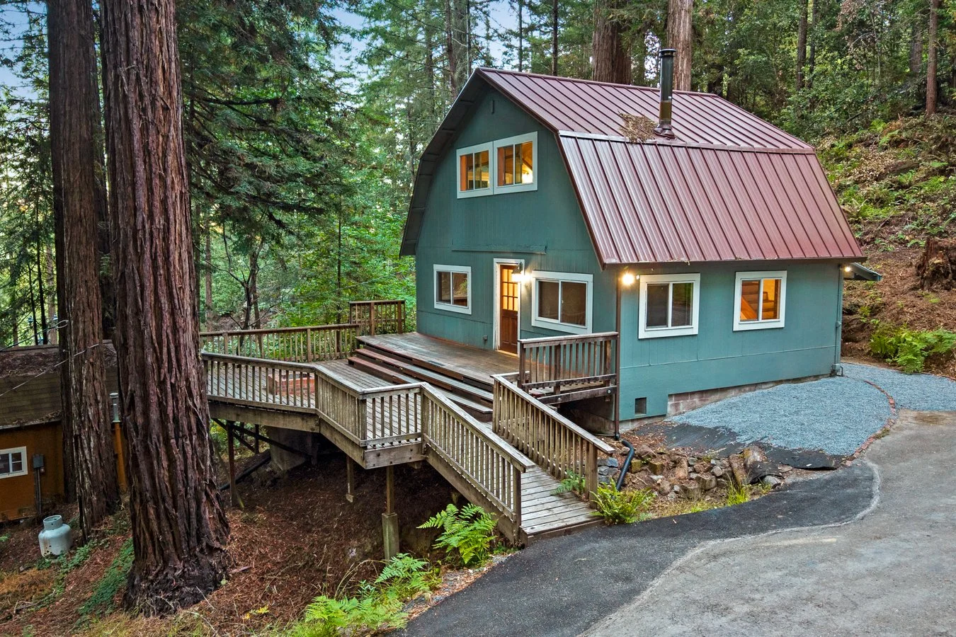 A two-story blue house with a metal roof, surrounded by tall trees, with a wooden deck and stairs leading to a gravel and asphalt driveway.