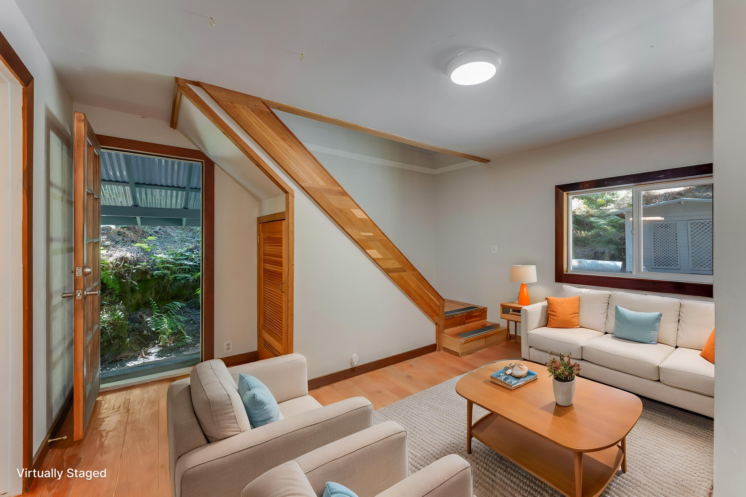 Living room with white couches, a coffee table, large window, open door showing greenery outside, wooden stairs, and a ceiling light.