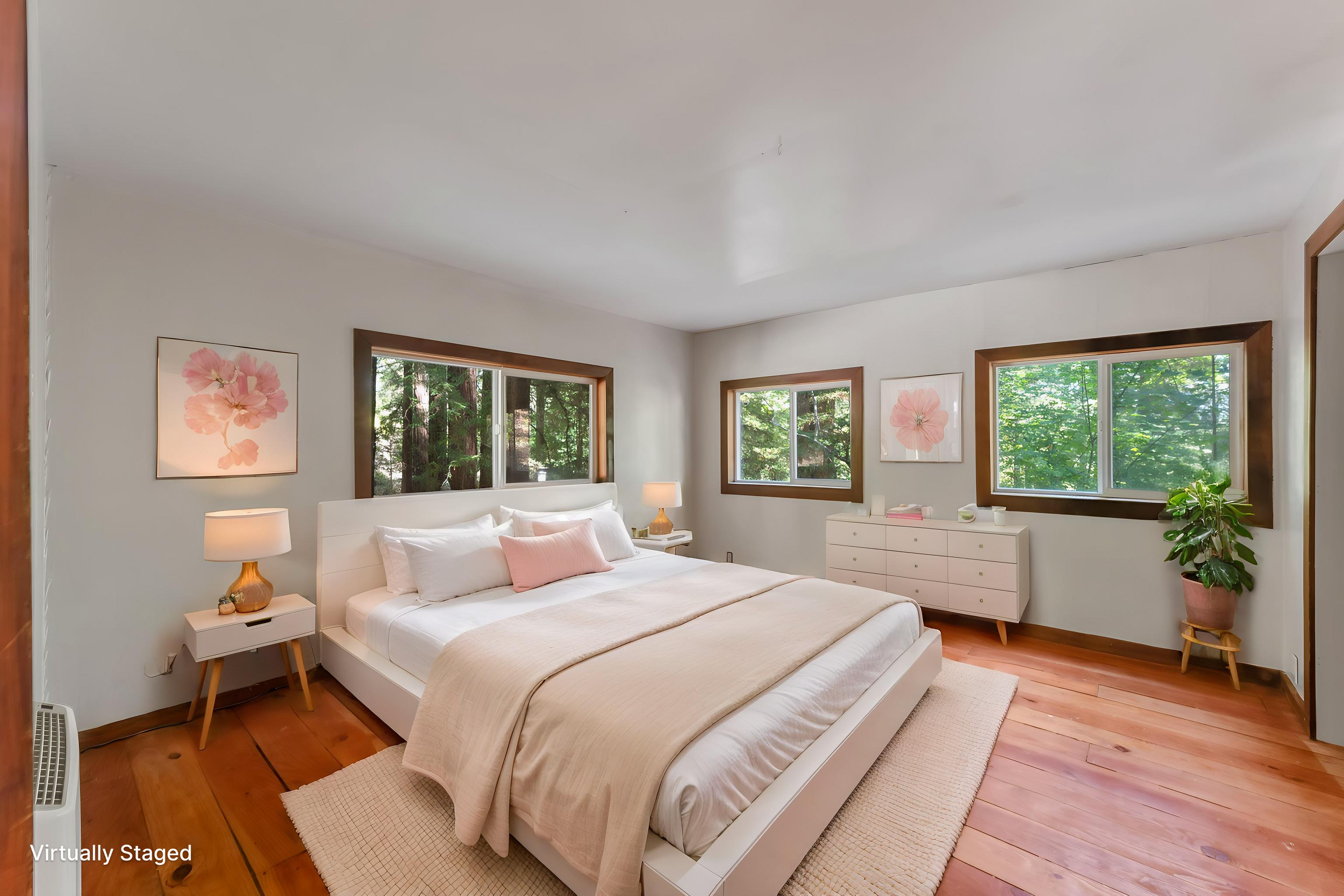 A bedroom featuring a large white bed with white, pink, and beige bedding, two nightstands with lamps, framed floral artwork, three windows showing greenery outside, a white dresser, and a potted plant, with hardwood floors and a beige rug.