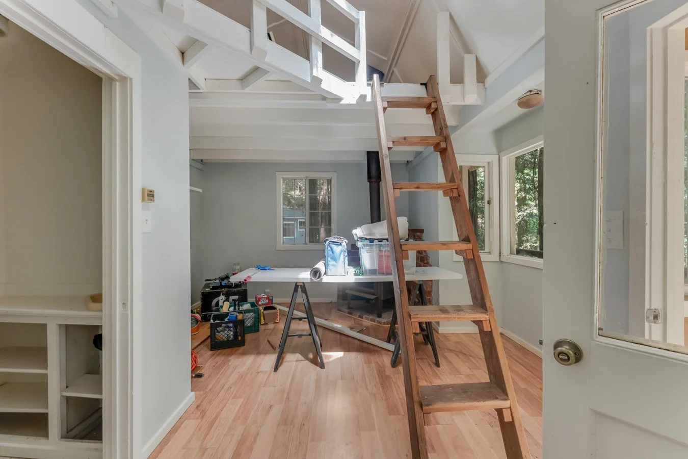 Interior of a house under renovation with a wooden ladder, tools, and construction materials in a room with large windows and a view of trees outside.