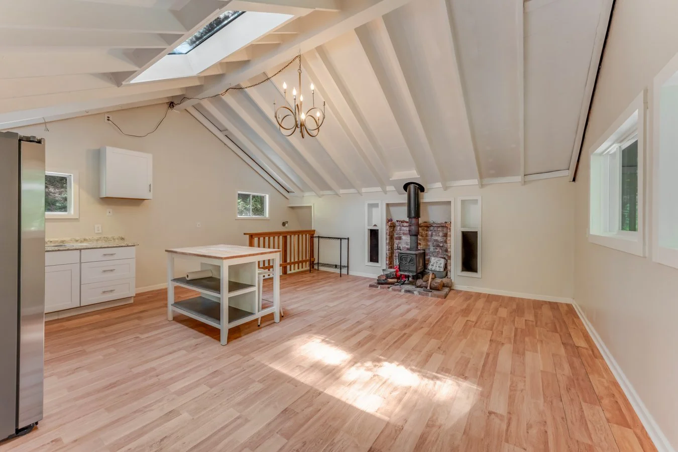 Empty attic room with wood flooring, white walls, windows, a wood-burning stove with brick surround, a chandelier, and open ceiling beams.
