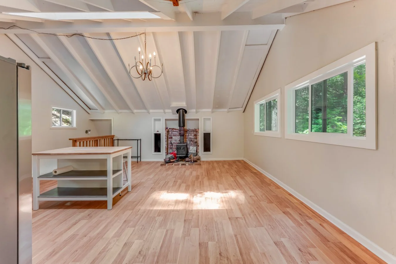 Empty living area with wooden floors, a brick fireplace, three large windows showing greenery outside, a chandelier, and a kitchen island with shelving.