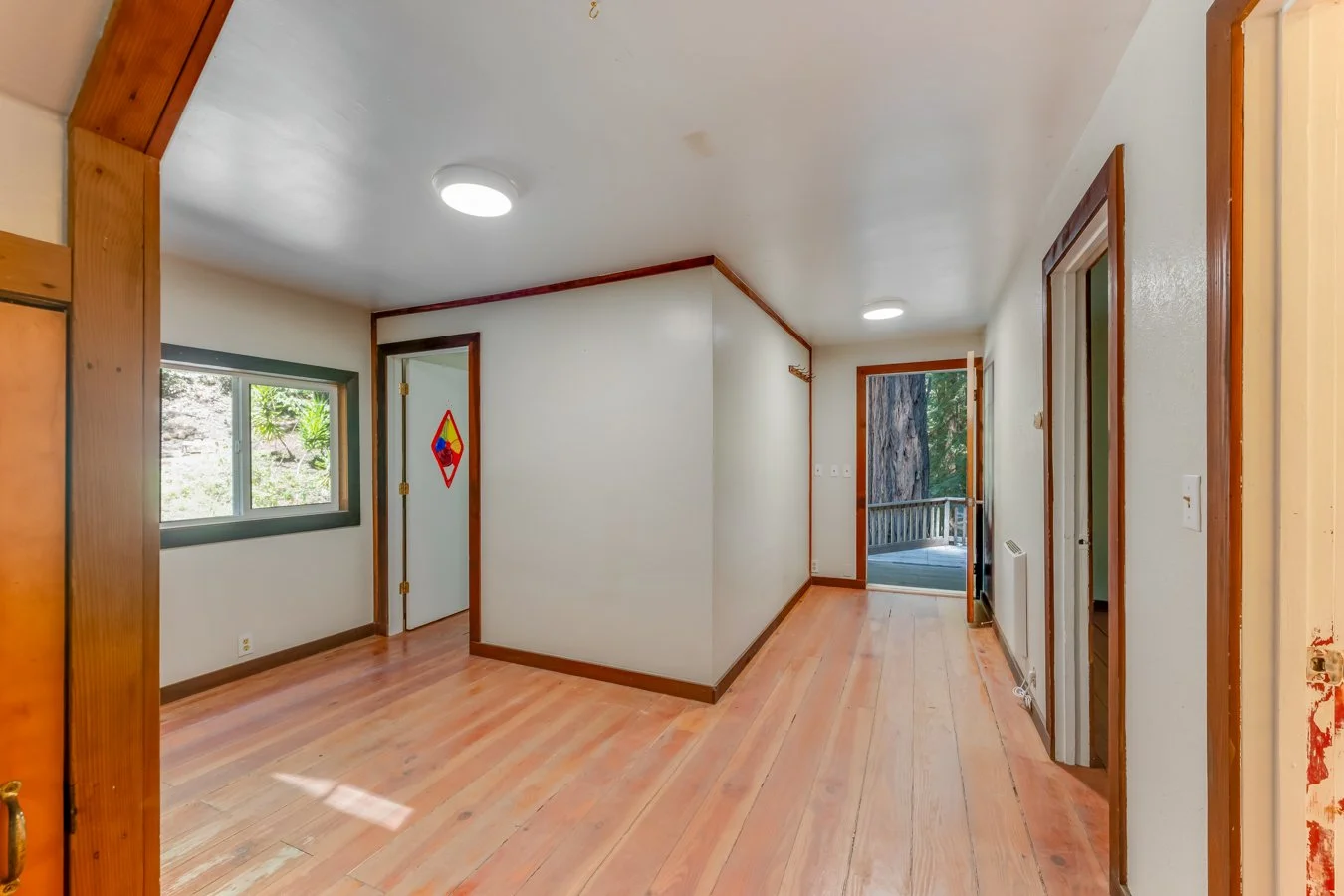 Empty room with wooden floors, white walls, and wooden trim. There are two sliding glass doors leading to a balcony with trees outside, and a window on the left wall. The room is lit with ceiling lights.
