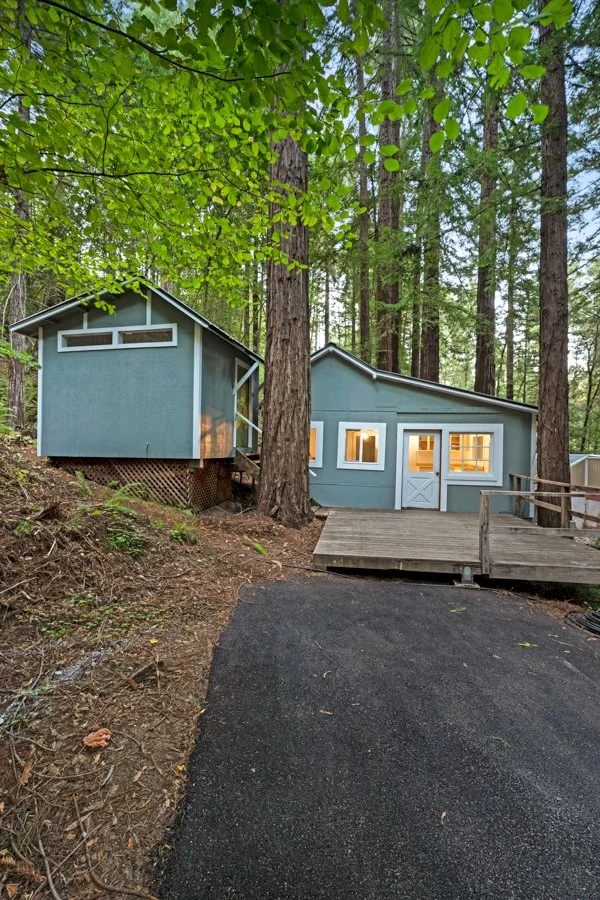 A small house in a forested area with tall trees, featuring a light blue exterior, a wooden ramp leading up to the entrance, and warm interior lighting visible through the windows.