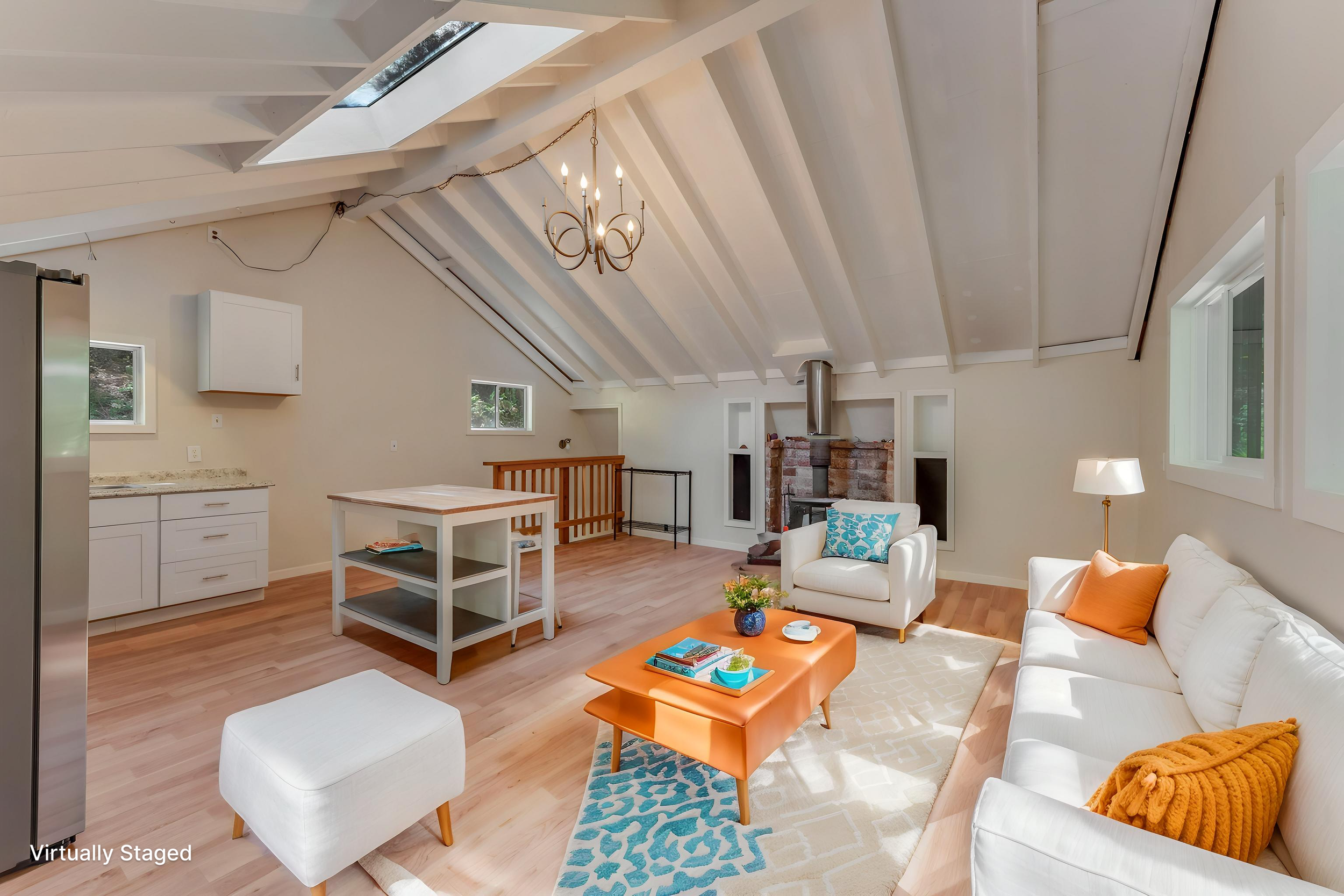 A spacious living room with vaulted ceiling, skylight, and wooden flooring. It features white sofas with orange and blue cushions, a small white coffee table, a patterned rug, a brick fireplace, and large windows bringing in natural light.