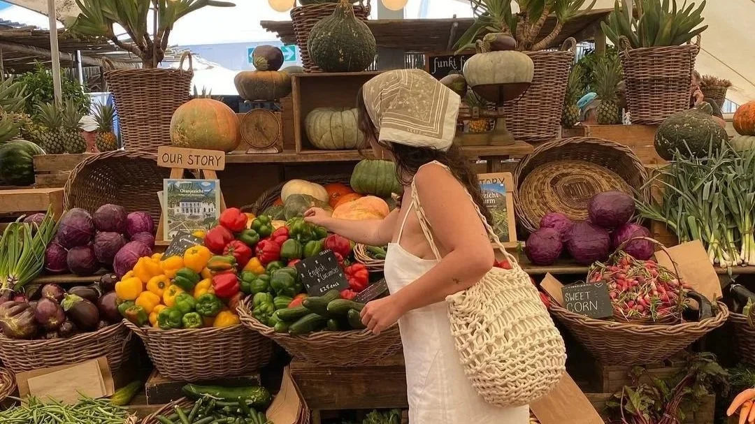A woman selecting vegetables at a farmers market stall with baskets of colorful peppers, pumpkins, and other produce.