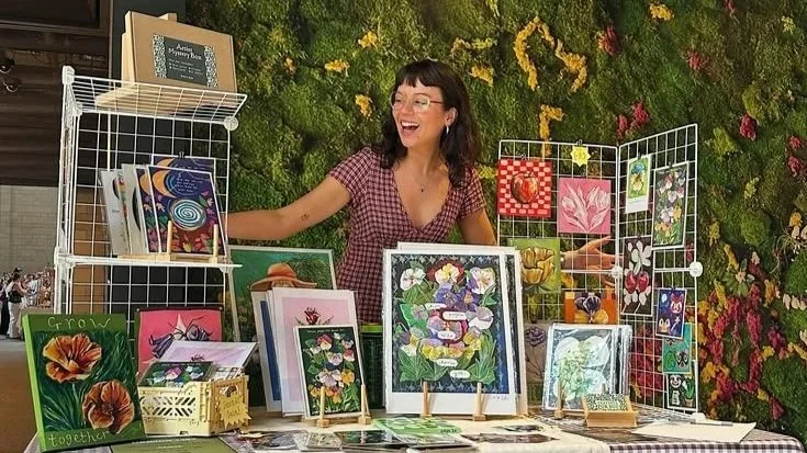 A woman with dark hair, glasses, and a red checkered shirt is smiling at a booth displaying colorful artwork and greeting cards, with a green plant wall in the background.
