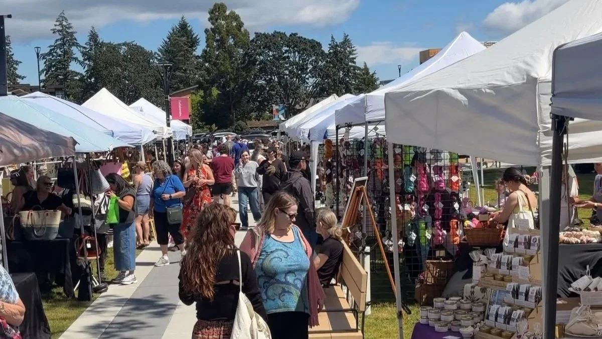 People browsing outdoor market stalls with white tents, selling various goods on a sunny day with trees and blue sky in the background.