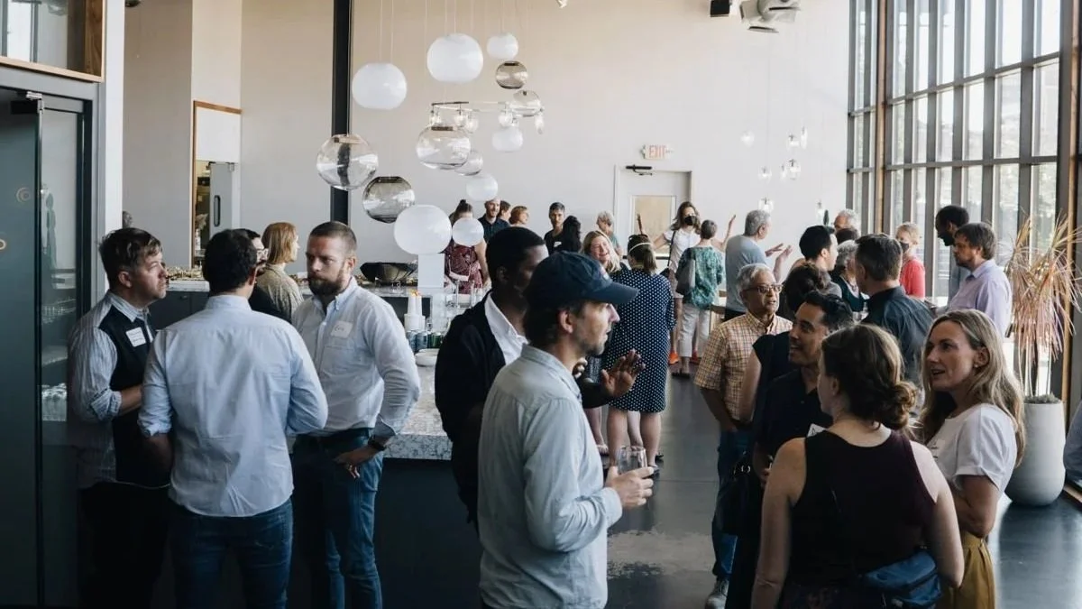 Group of people socializing at an indoor event with large windows, hanging pendant lights, and a table with food and drinks.
