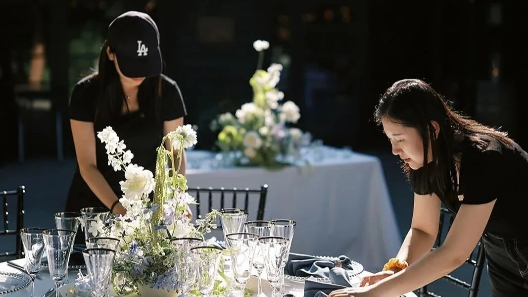 Two women setting a table with a floral centerpiece at an outdoor event