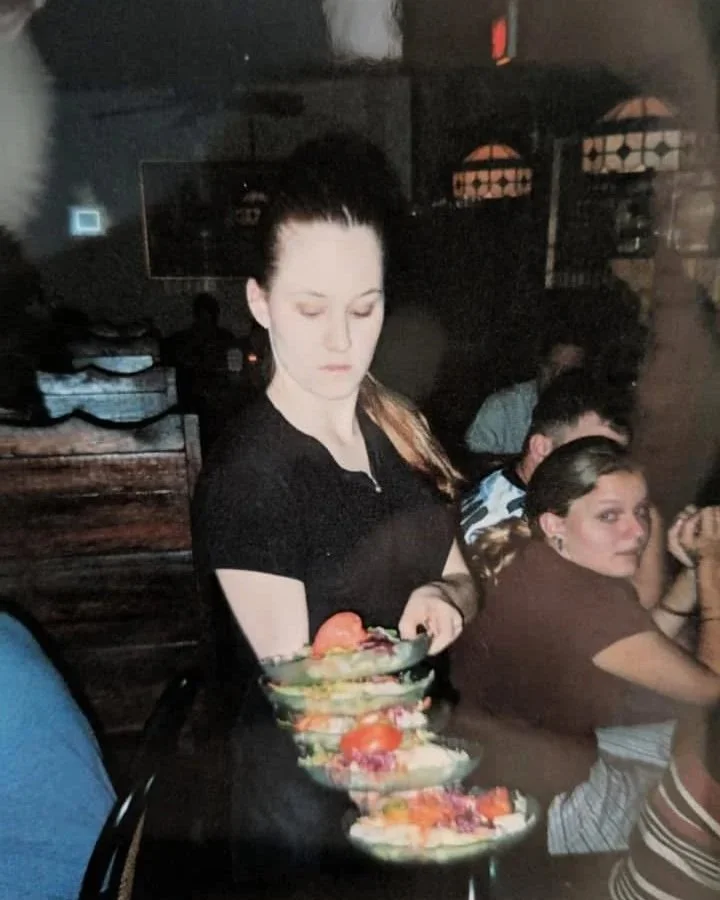 A woman in a black shirt is holding a three-tiered serving tray with desserts in a dimly lit restaurant. Several people are seated at tables in the background.