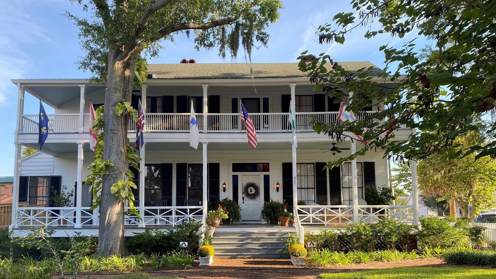 Front view of a large white house with two stories, multiple black shutters, and a porch with white columns. Several flags hang from the upper porch, including American and state flags. There are stairs leading up to the front door, which is decorated with a wreath. The house is surrounded by greenery, including trees and bushes, and potted plants are positioned near the stairs.