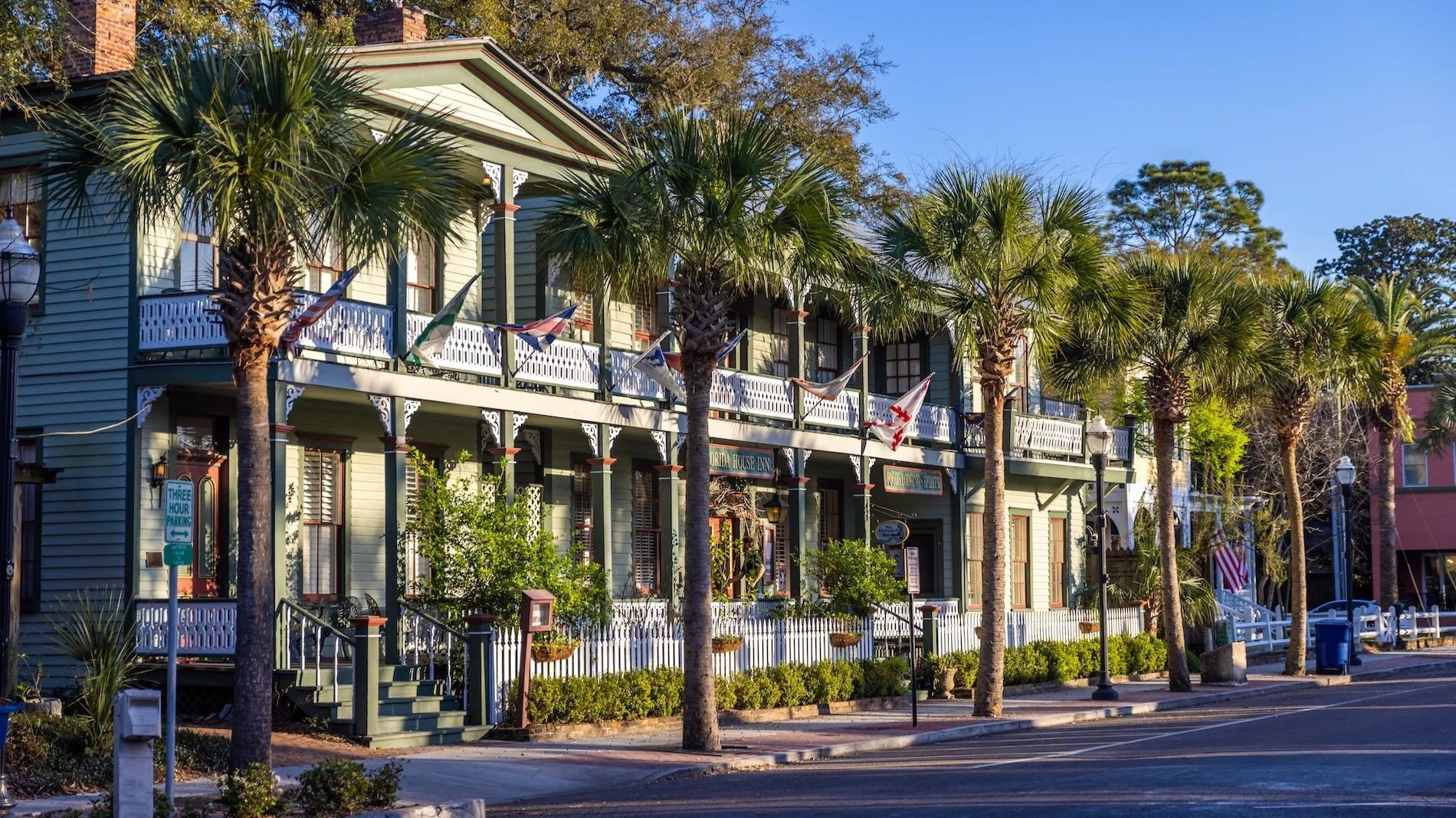 A row of wooden buildings painted in light green with white trim and balconies, with flaming torch lights and hanging flags. There are tall palm trees in front of the buildings and a clear blue sky in the background. A sign indicates a three-hour parking limit.