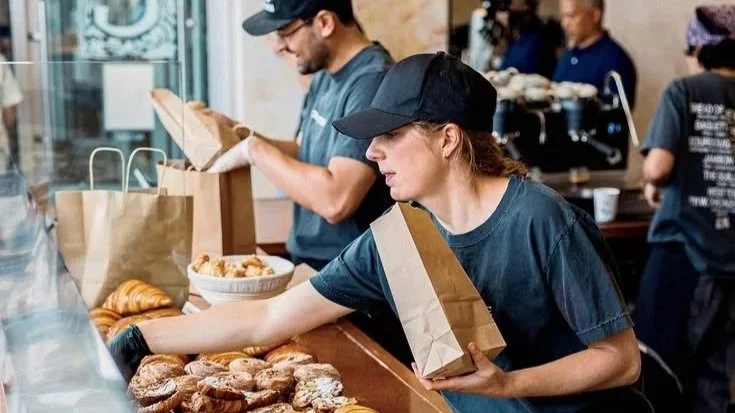 Two women working behind a bakery counter with baked goods, other staff in the background, in a busy bakery or cafe.
