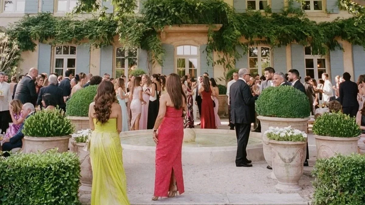 People dressed in formal attire at an outdoor event in a courtyard with a fountain, potted plants, and a building with window shutters in the background.