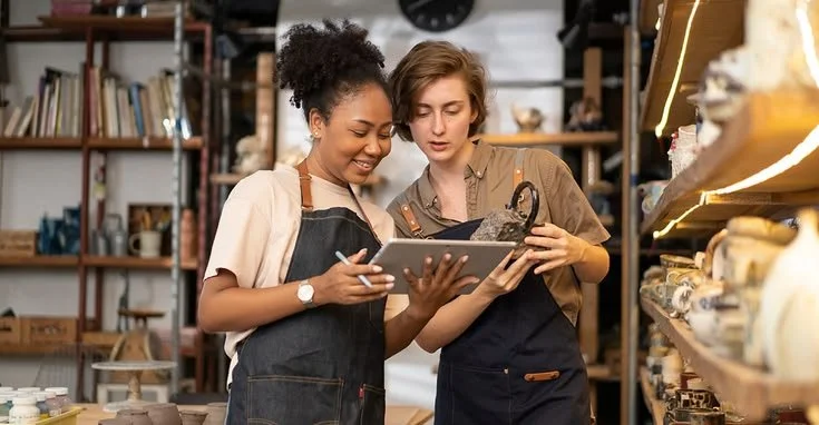 Two women in an art or craft store, one with curly hair and one with short hair, looking at a tablet and a ceramic item.