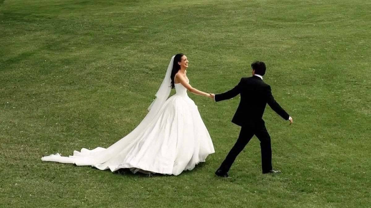 Bride in a white wedding dress and veil holding hands with groom in a black suit, on a grassy field.