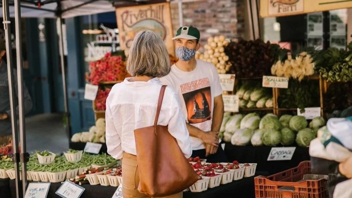 A woman with gray hair carrying a brown shoulder bag shopping at a farmer's market stall, talking to a young male vendor wearing a white T-shirt with a graphic print, a green and white cap, and a face mask, with fresh vegetables like strawberries, green beans, cabbage, and flowers displayed behind them.