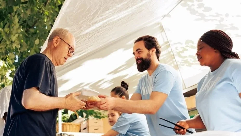 A group of people, including an elderly man receiving food, are outdoors under a canopy. Two women and a man wearing casual light blue T-shirts assist with serving food at what appears to be a community event or charity. The setting is sunny with green foliage in the background.