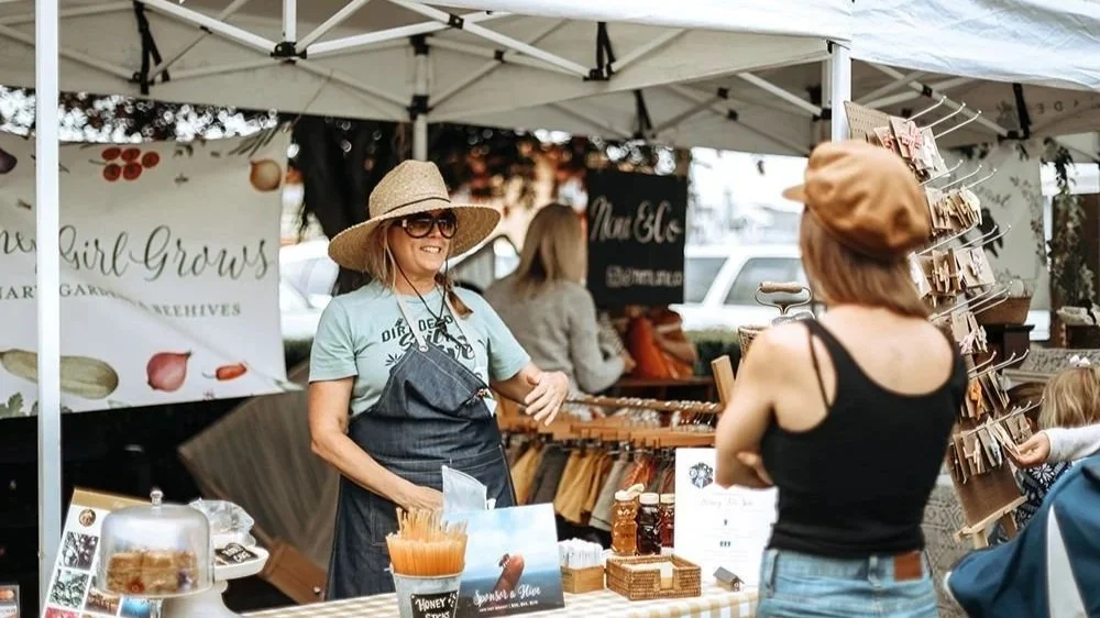 A woman selling honey and bee-themed products at an outdoor market stall, talking to a customer in a black tank top and brown hat.