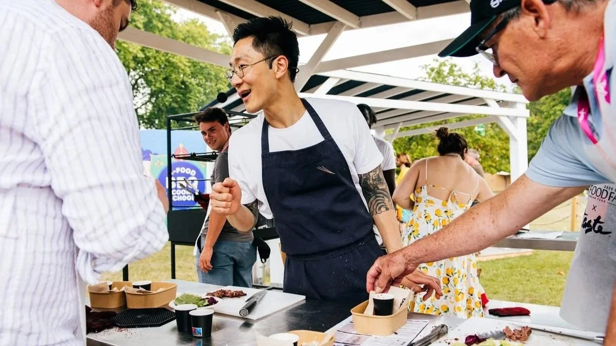 People participating in a food-tasting event outdoors, with a chef holding a microphone and engaging with visitors at a booth.