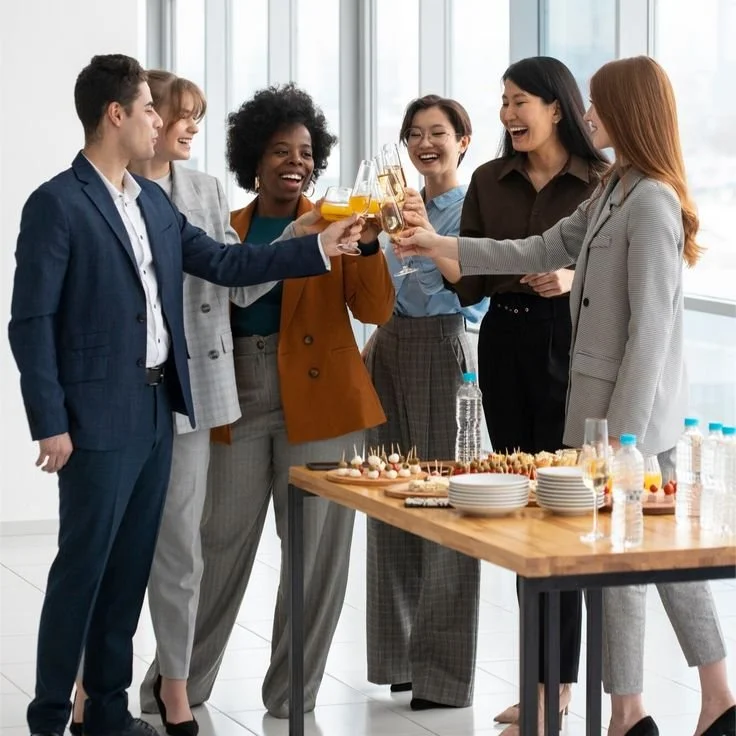 Group of six diverse businesswomen and businessmen celebrating at a professional gathering with drinks and snacks.