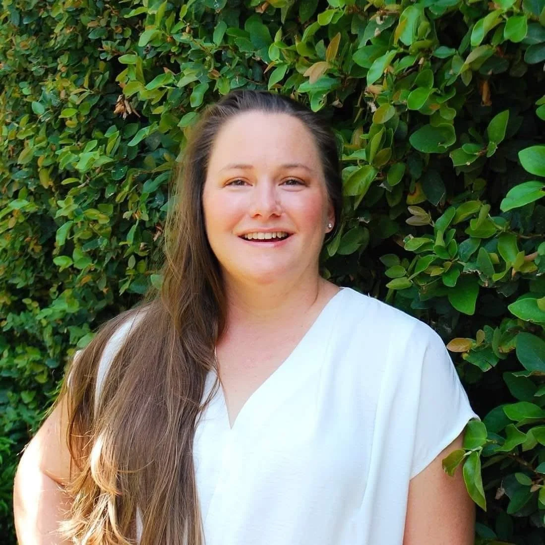 A young woman with long brown hair and a white top standing in front of a green leafy bush, smiling at the camera.