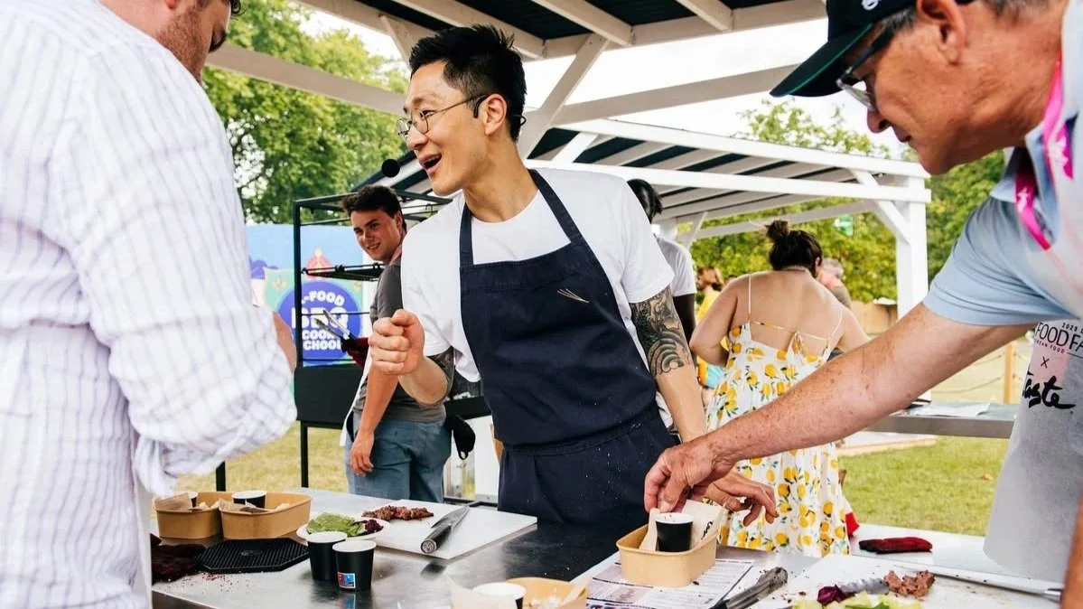 A man with glasses and tattoos wearing a white t-shirt and black apron is talking and laughing with two men at an outdoor food event. One man is in a white striped shirt, and the other is wearing a light blue shirt and cap. There are food samples on the table and a food festival banner in the background.