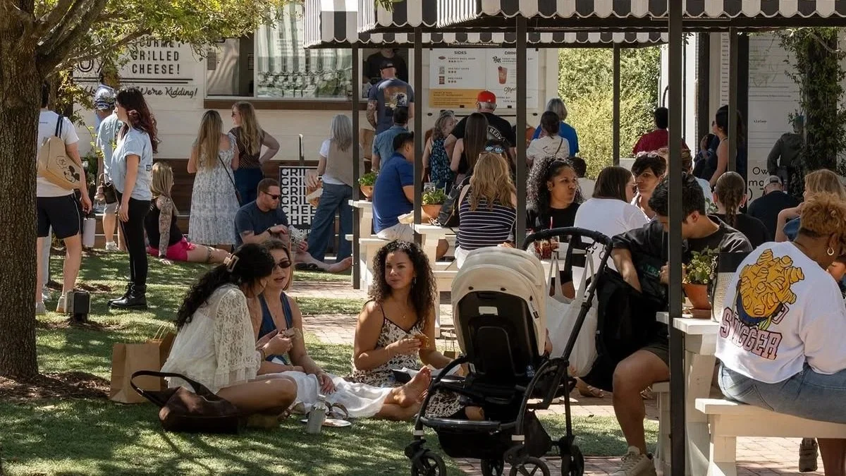 Outdoor scene with many people gathered around a food truck and seated at tables, some standing in line, enjoying food and conversation under a tree and umbrellas on a sunny day.