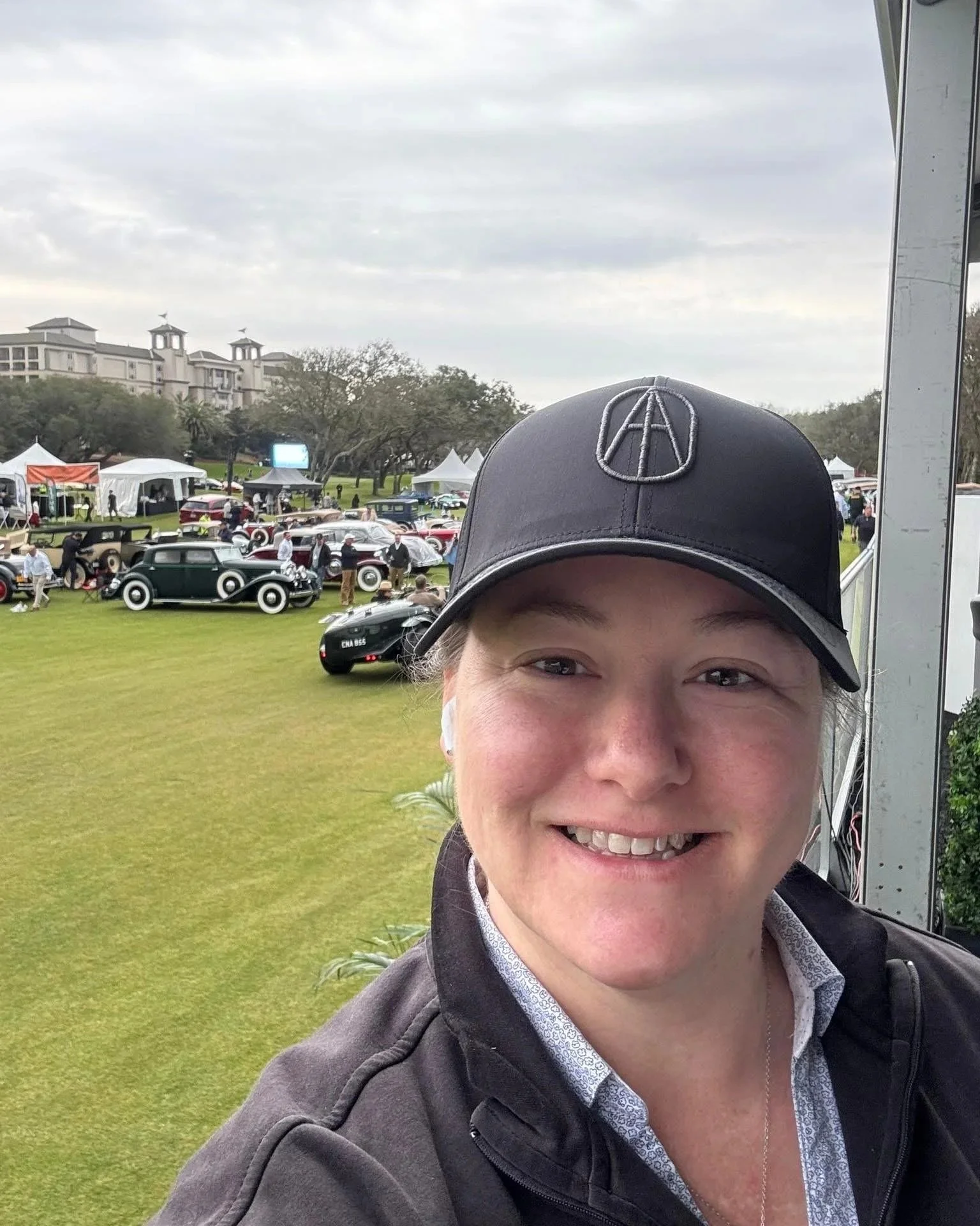A person smiling outdoors at a classic car show, wearing a black cap with an 'A' emblem, with vintage cars and tents in the background.