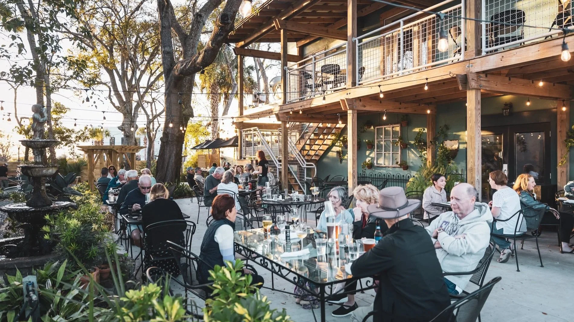 People dining outside at a restaurant patio during sunset, with a two-story wooden building and trees in the background, string lights hanging overhead.