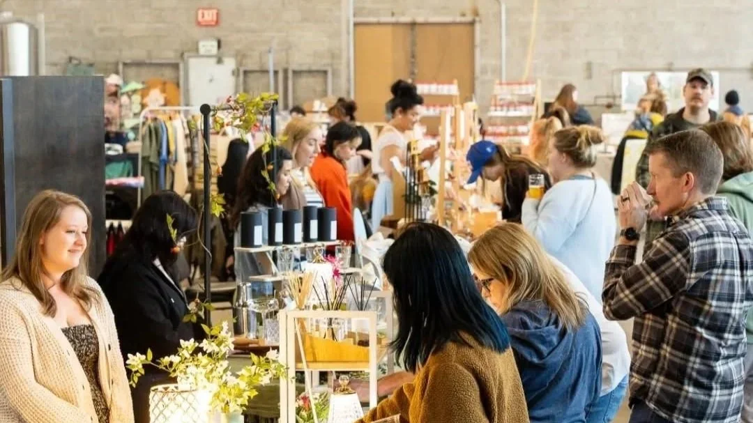 People shopping at a craft fair or market, browsing stalls with handmade items in an indoor warehouse setting.
