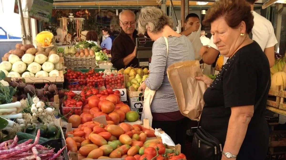 People shopping at a farmers market with a table of fresh vegetables and fruits, including tomatoes, onions, and apples.