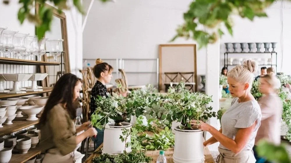 A group of women arranging green plants on a long wooden table indoors, with shelves of white dishes and glasses in the background.