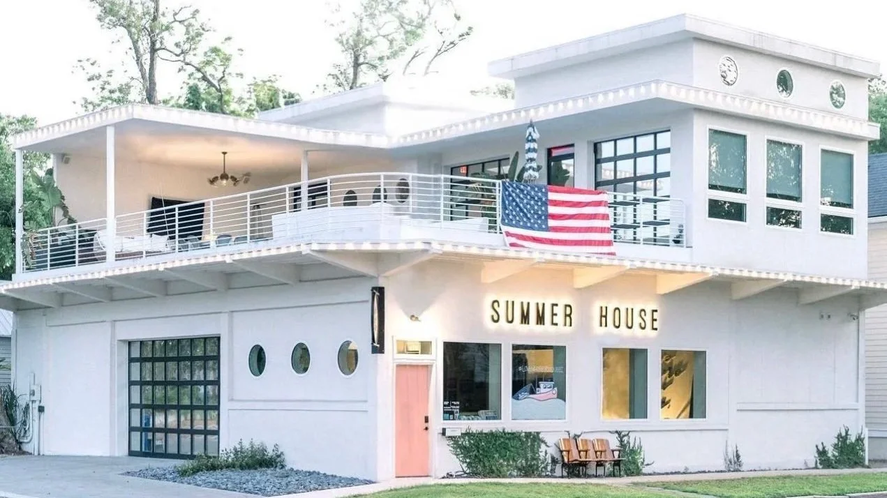 A white two-story house with a sign that reads 'Summer House' on the front. The house has a balcony with outdoor seating and an American flag hanging from it. There are trees in the background and a small lawn in front.