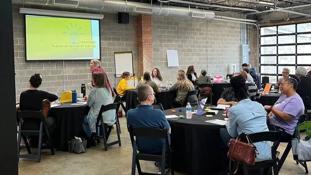 People attending a workshop or seminar in a spacious industrial-style room with large windows, black tables, and a projection screen showing a welcome message.