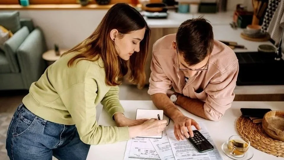 A young woman and a man are reviewing documents on a kitchen counter. The woman is writing in a notepad, and the man is using a calculator. The kitchen has a modern style with shelves, plates, and a cup of tea.