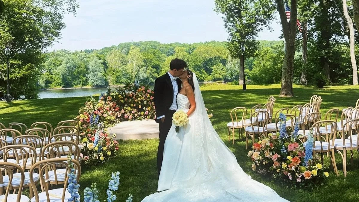 A bride and groom kiss during an outdoor wedding ceremony, with chairs and floral arrangements in a lush green park.