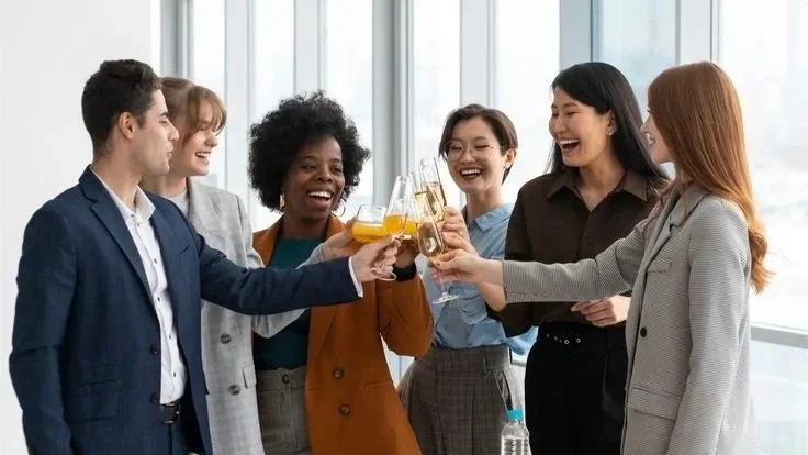 Six diverse young adults in business attire celebrating together with drinks in a bright office.