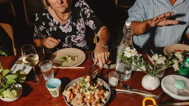 A woman serving food at a dinner table, with dishes, drinks, and decorative flowers.