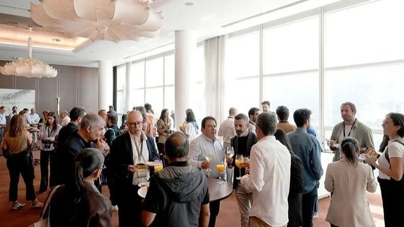 People socializing and having drinks at a modern indoor conference or networking event with large windows, white walls, and ceiling decor.