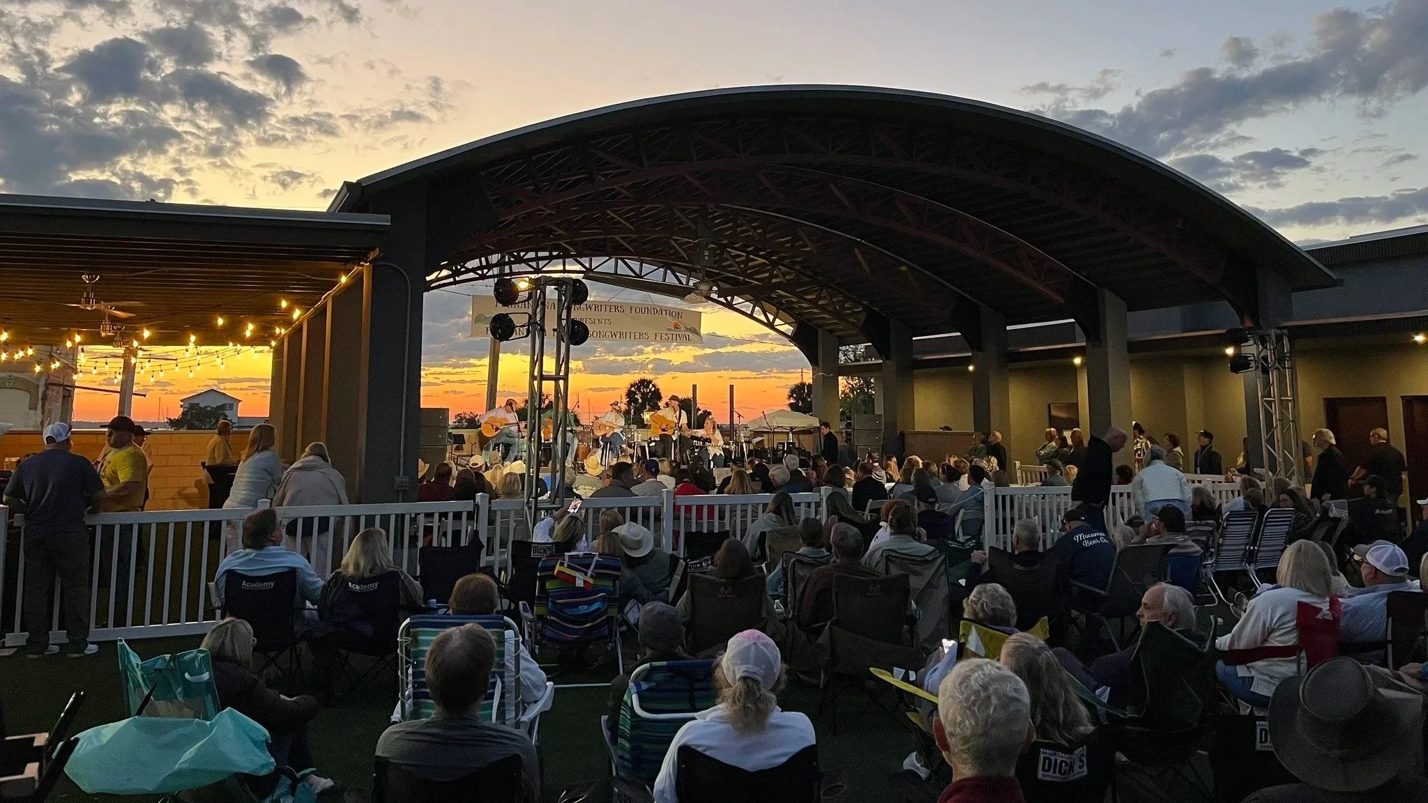 An outdoor concert stage with musicians playing guitars at sunset, surrounded by seated audience and string lights.