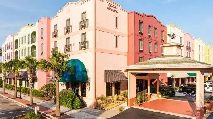 Colorful multi-story buildings with palm trees and a parking lot, featuring a pink and beige color scheme and a covered entrance.