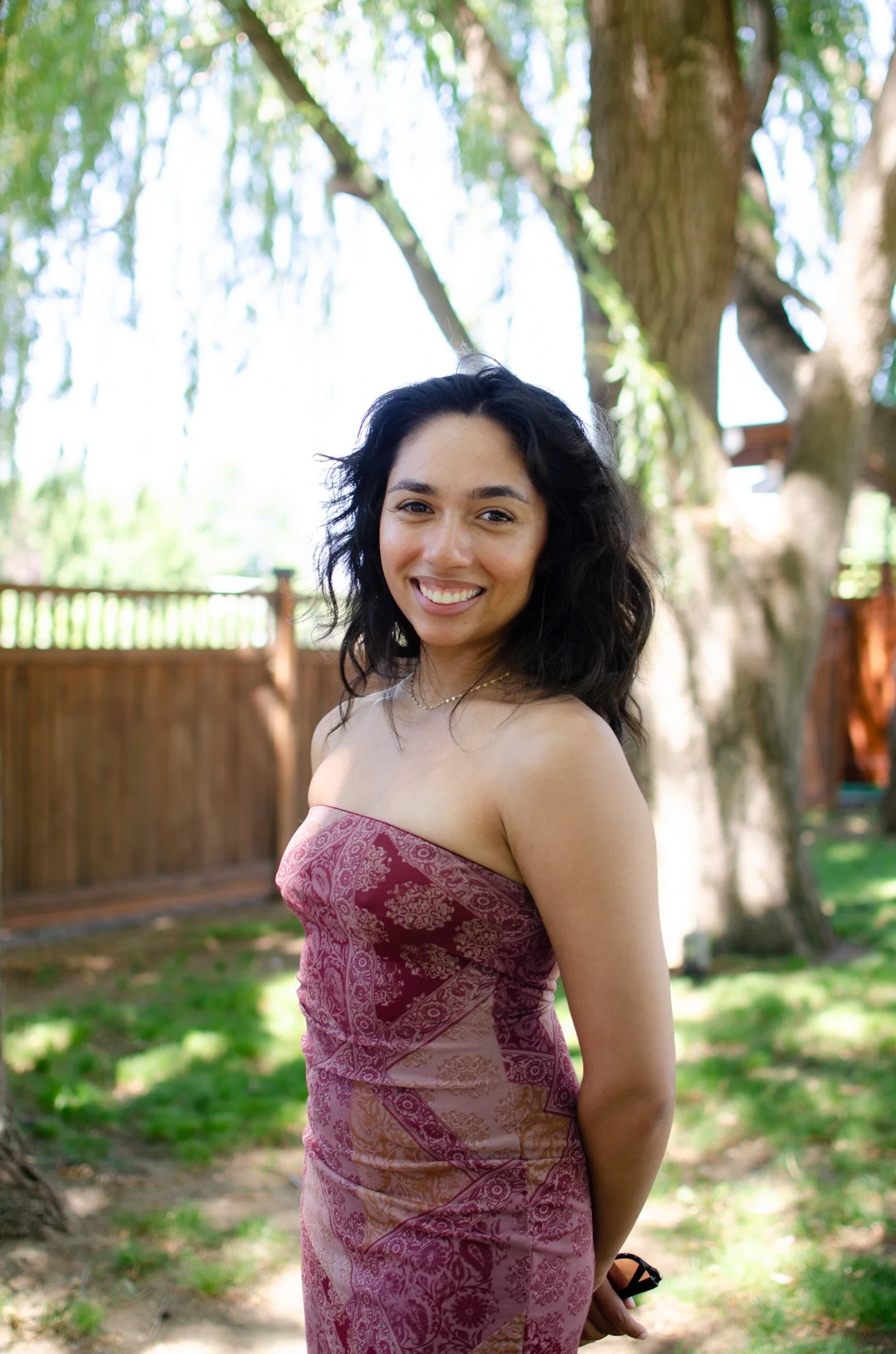 A woman with long, curly black hair smiling outdoors, wearing a strapless, patterned pink and purple dress, with a wooden fence and green trees in the background.