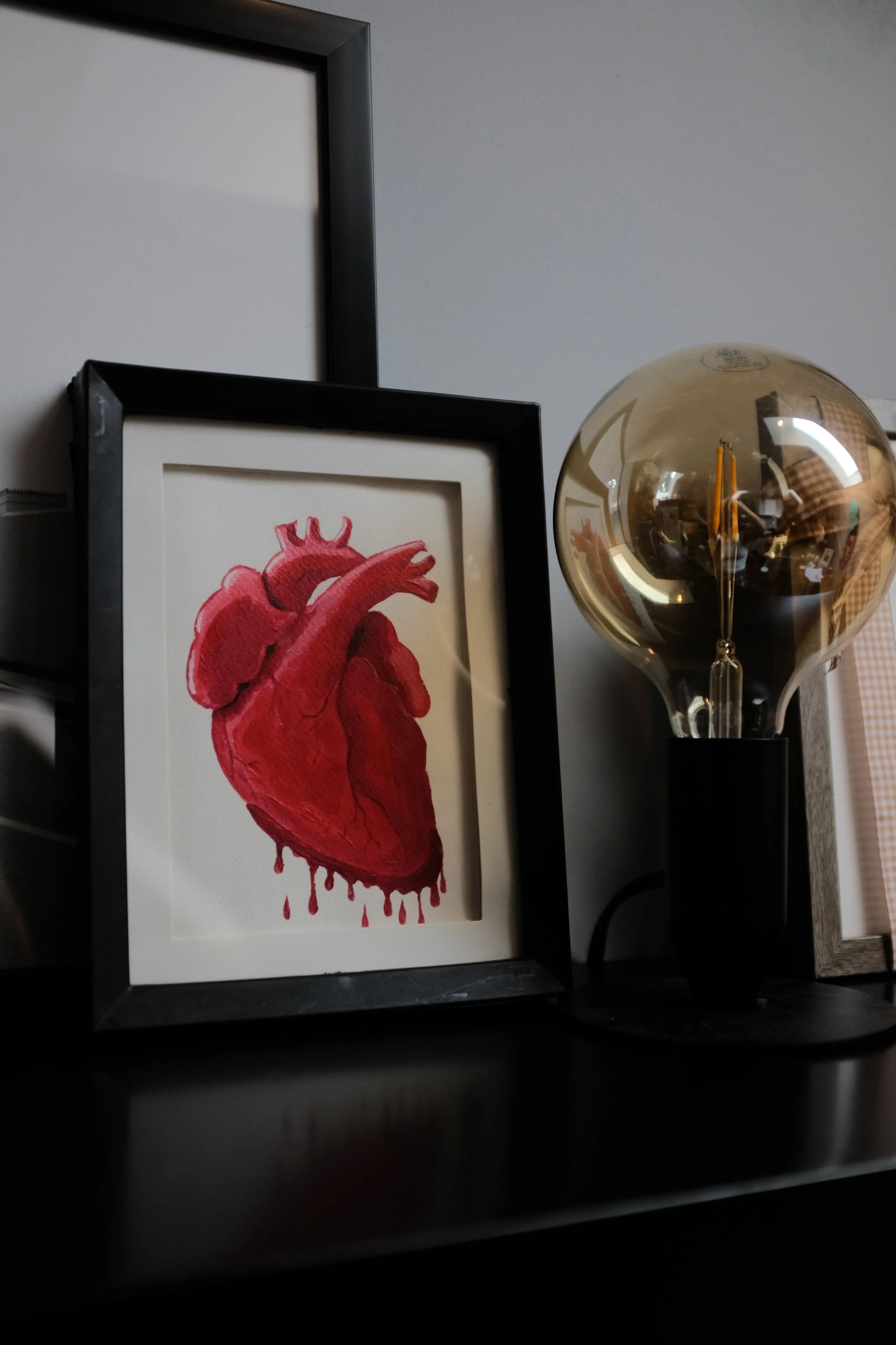 A framed watercolor painting of a human heart with blood dripping from the bottom, leaning against a black surface. Partially visible framed artwork and a reflective gold spherical lamp are in the background.