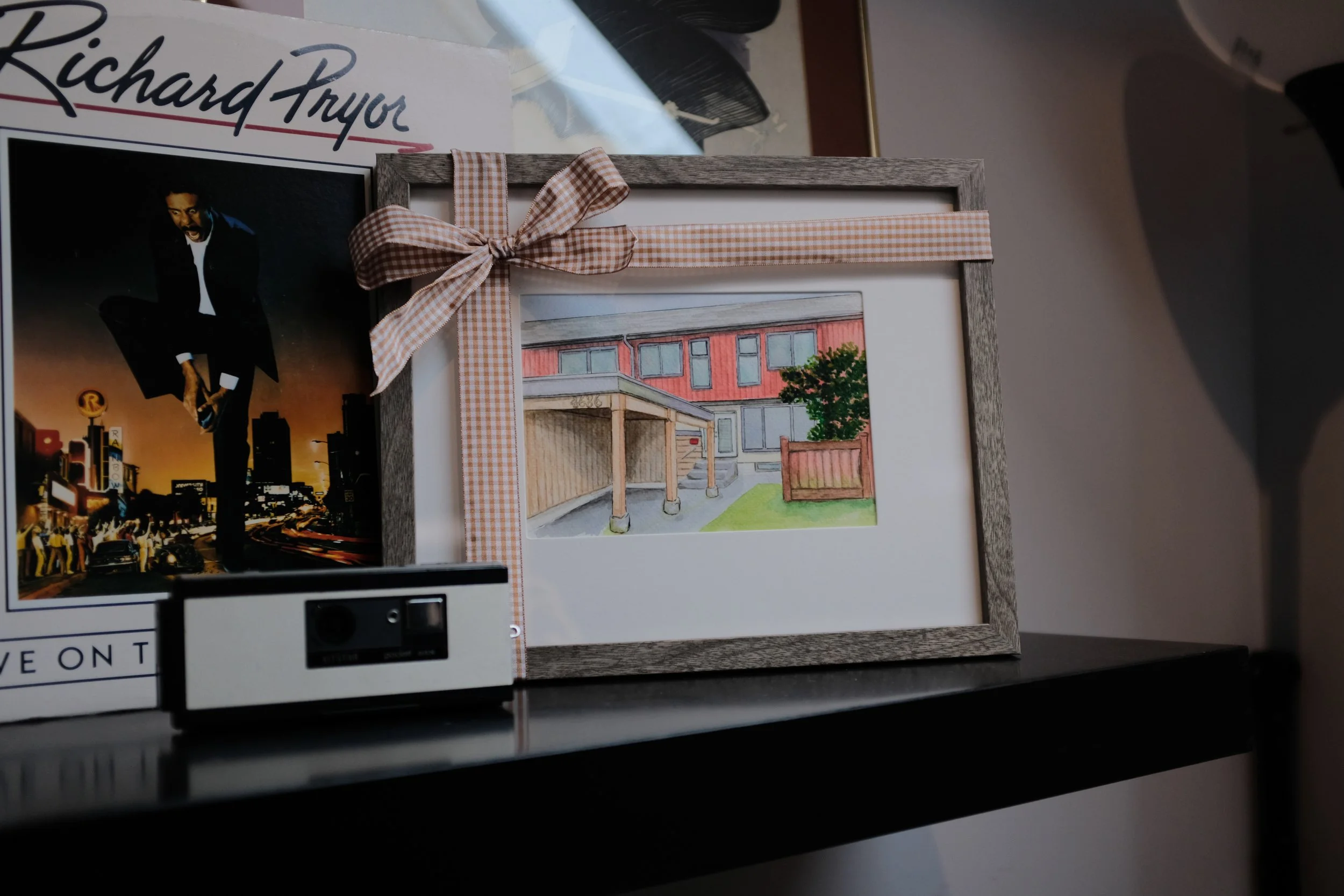 Photograph of a framed picture of a house, a colorful cityscape poster, and a photograph of a man in black attire with an urban background, all resting on a white surface.