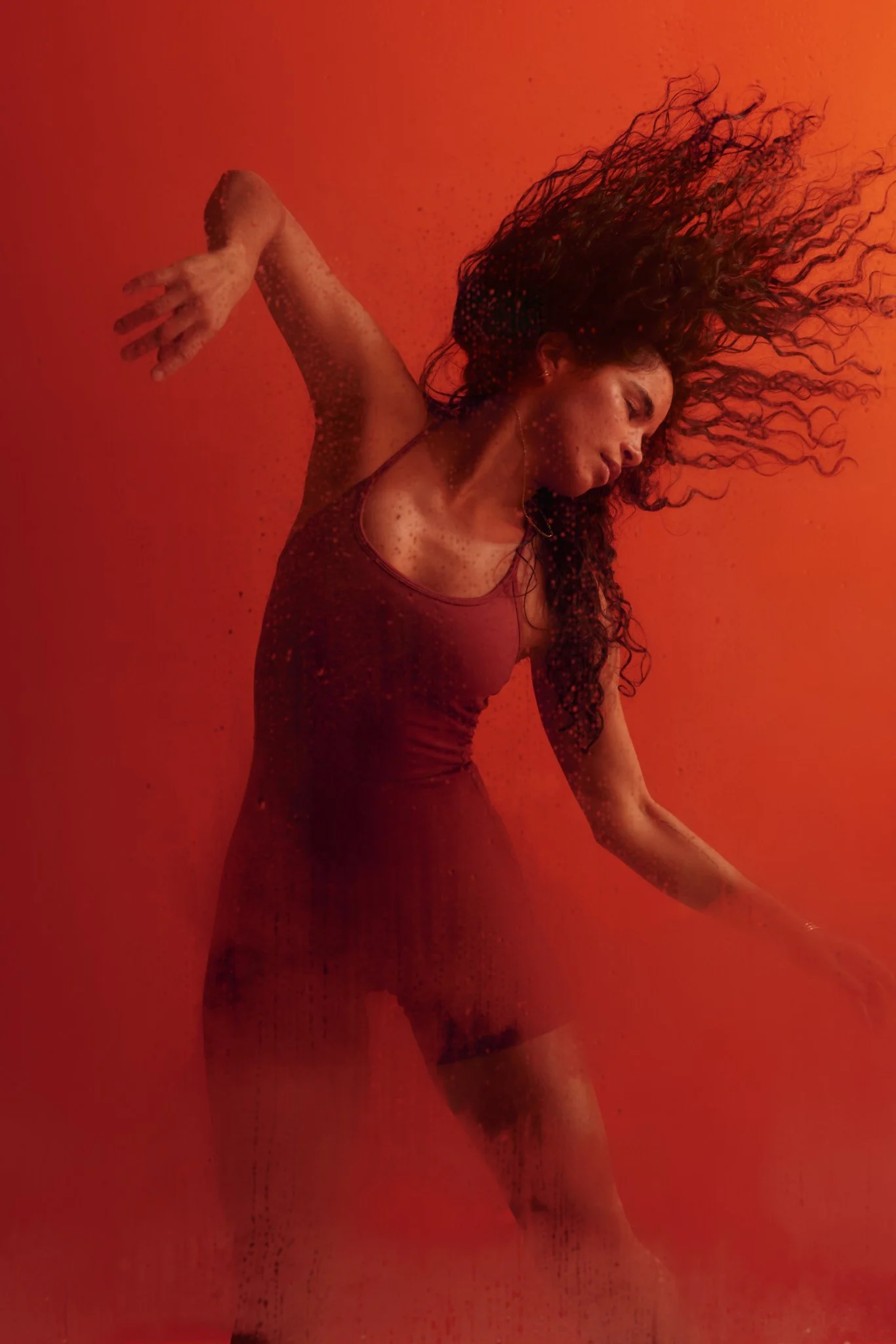 Woman with long curly hair posing in a red-lit studio, wearing a dark dress with her arms outstretched