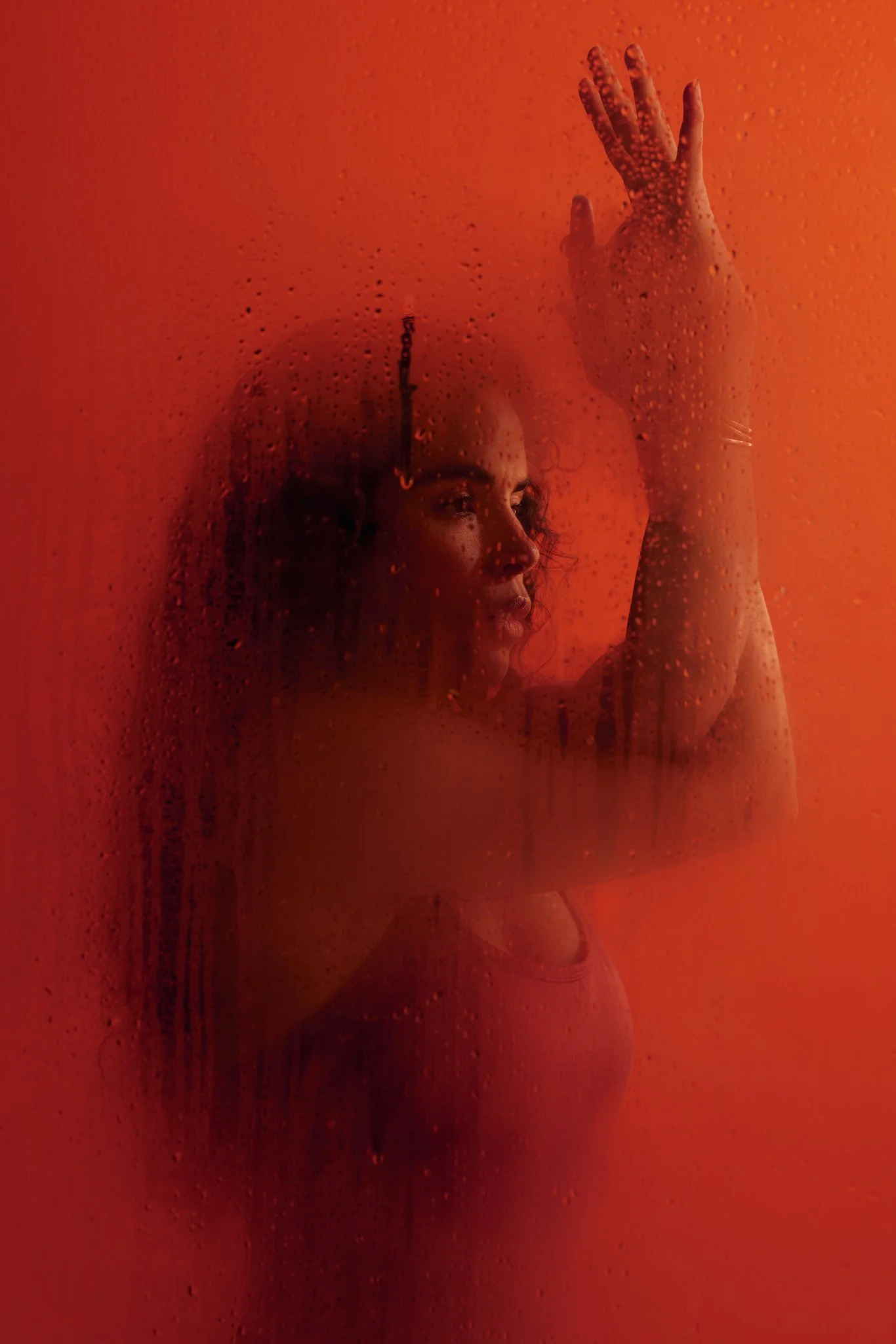 Woman seen through a rain-covered glass window, raising her hand in dramatic red-orange lighting